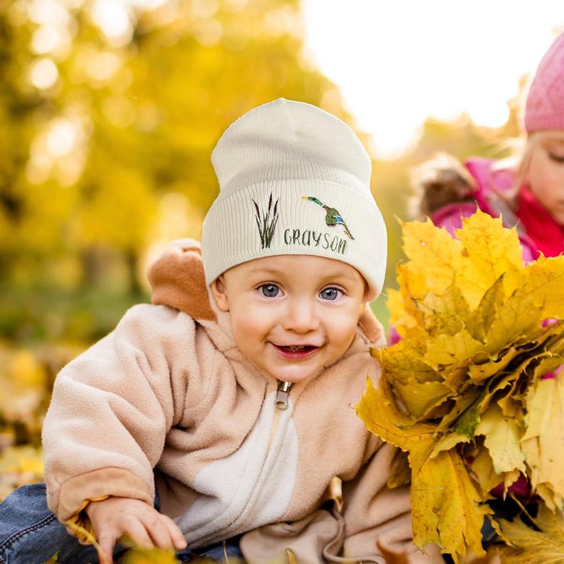 May include: A cream-colored beanie with the name "Grayson" embroidered in a script font, along with a duck and cattails design. The beanie is worn by a child. The child is wearing a tan jacket and is surrounded by yellow leaves.