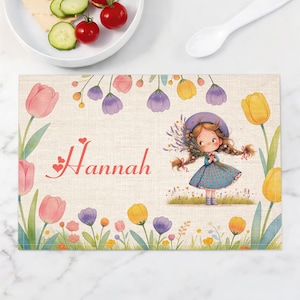 May include: A rectangular placemat with a floral design and the name "Hannah" in red script. The placemat features a cartoon girl holding flowers, surrounded by pink, purple, and yellow tulips. The background has a linen texture.