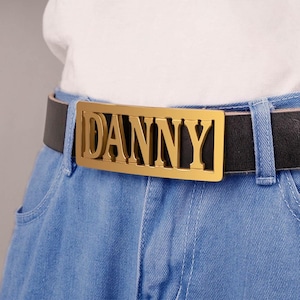 May include: A black leather belt with a gold-coloured buckle spelling "DANNY". The belt is worn with blue jeans and a white shirt. The buckle is rectangular and the name is in capital letters.
