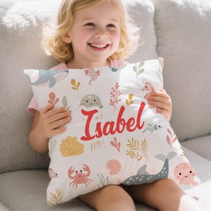 Könnte beinhalten: Quadratisches weißes Kissen mit einem Meeresmotiv, das Meeresbewohner und Pflanzen zeigt. Das Kissen hat ein Wal-, Seepferdchen-, Krebs- und Oktopus-Design. Der Name "Isabel" ist in roter Schrift über der Mitte des Kissens geschrieben.