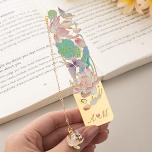 May include: A gold-toned metal bookmark featuring an intricate cutout design of lotus flowers and butterflies with pastel pink, blue, and green enamel accents. A delicate gold chain hangs from the top, ending in a small lotus charm. The base is engraved with the letters A and M separated by a heart symbol.