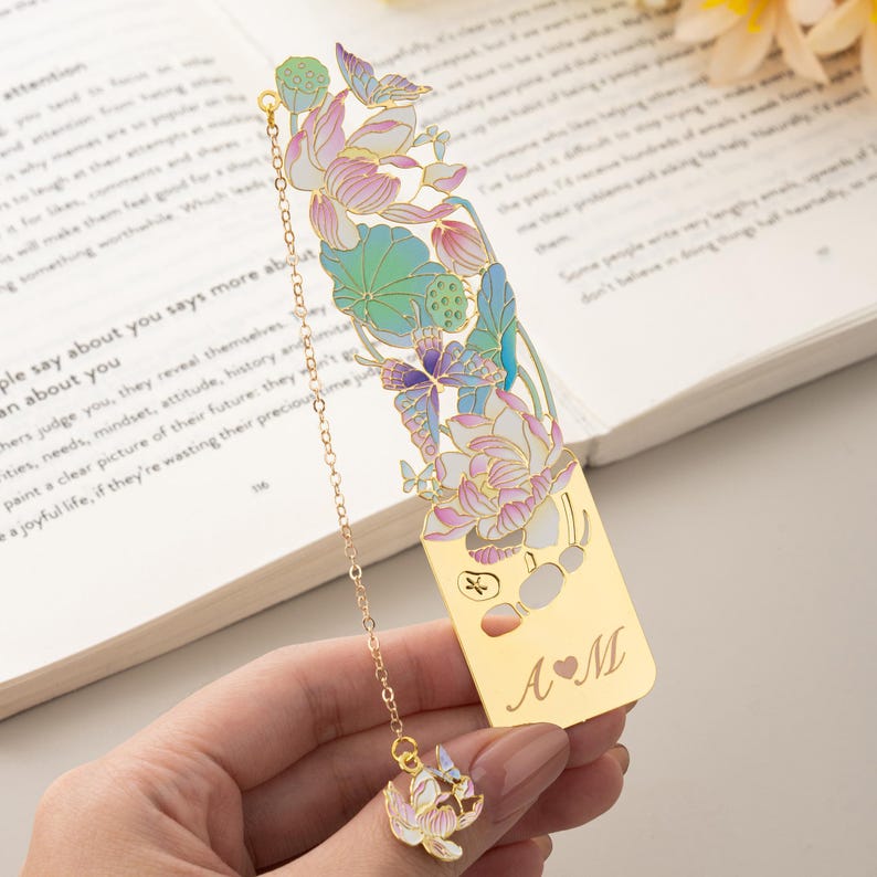 May include: A gold-toned metal bookmark featuring an intricate cutout design of lotus flowers and butterflies with pastel pink, blue, and green enamel accents. A delicate gold chain hangs from the top, ending in a small lotus charm. The base is engraved with the letters A and M separated by a heart symbol.
