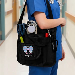 Puede incluir: Un bolso organizador médico de nailon negro con correa ajustable para el hombro, que cuenta con múltiples bolsillos exteriores llenos de herramientas de enfermería. El bolsillo frontal muestra un lazo bordado con el texto NICU y el nombre Carolyn, RN.