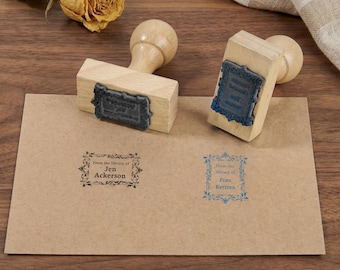 May include: Two wooden stamps with text and decorative borders sit on a brown envelope. The stamps have a rectangular design with a wooden handle. The envelope has two stamped impressions, one black and one blue, with the text "From the library of" and a name.