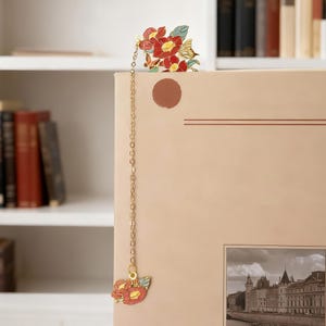 May include: A metal bookmark featuring a gold-toned chain connecting two floral charms. The top charm displays red camellia flowers with green leaves, while the bottom charm features a smaller matching floral design in red and light blue enamel.