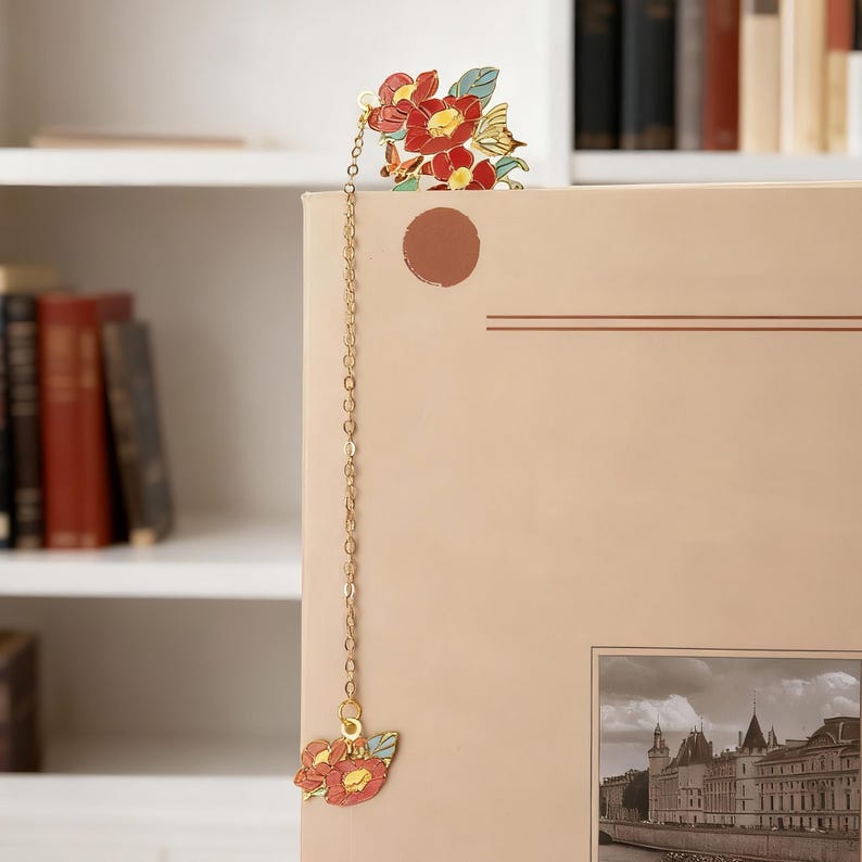 May include: A metal bookmark featuring a gold-toned chain connecting two floral charms. The top charm displays red camellia flowers with green leaves, while the bottom charm features a smaller matching floral design in red and light blue enamel.