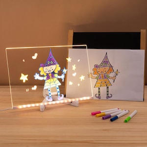 May include: A light-up drawing board featuring a cartoon witch illustration. The board is illuminated with warm white lights, and the drawing is replicated on a nearby sheet of paper. Several colourful markers are visible on the wooden surface.