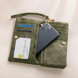 K&ouml;nnte beinhalten: Offene olivgr&uuml;ne Geldb&ouml;rse mit mehreren Kartenf&auml;chern, einem transparenten Ausweisfach und einem Smartphone. Die Geldb&ouml;rse hat eine Handschlaufe und einen Druckknopfverschluss. Das Smartphone ist dunkelgrau.