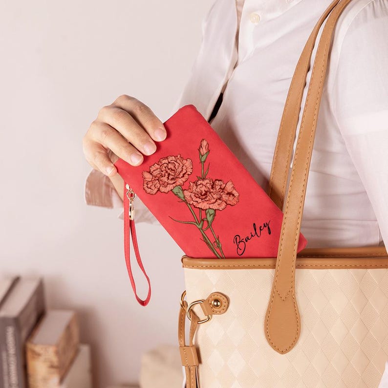 K&ouml;nnte beinhalten: Eine rote Geldb&ouml;rse mit floralem Design und dem Namen "Bailey" in Schreibschrift. Die Geldb&ouml;rse hat eine Handschlaufe und wird in der N&auml;he einer beigen Tragetasche mit braunen Riemen gehalten.