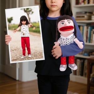 May include: A child holding a framed photograph and a puppet. The photograph shows a girl in a white shirt with black polka dots, red trousers, and white shoes. The puppet has a large red mouth, black hair, and is wearing a similar outfit.