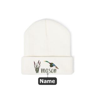 May include: A white beanie with a folded brim. The beanie features an embroidered design of a duck in flight, reeds, and the name "mason". Below the name is a black rectangle with the word "Name".