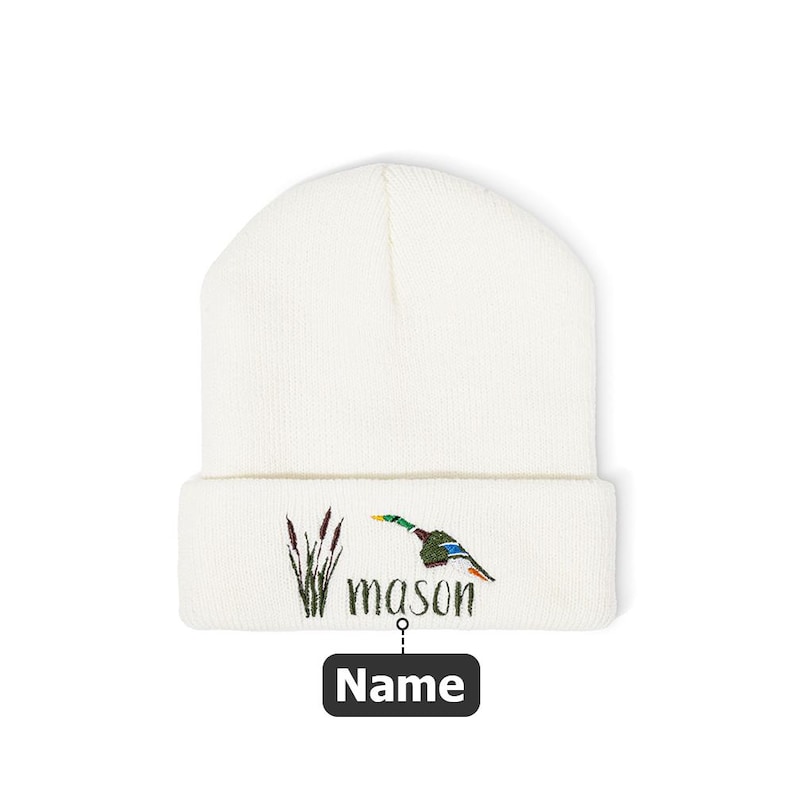 May include: A white beanie with a folded brim. The beanie features an embroidered design of a duck in flight, reeds, and the name "mason". Below the name is a black rectangle with the word "Name".
