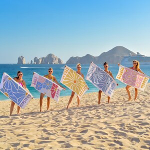 May include: Five women on a beach hold up personalized beach towels. Each towel features a floral design in different colors, with names printed on them. The background includes the ocean, mountains, and a cruise ship under a clear blue sky.