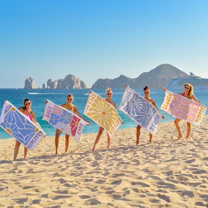 May include: Five women on a beach hold up personalized beach towels. Each towel features a floral design in different colors, with names printed on them. The background includes the ocean, mountains, and a cruise ship under a clear blue sky.
