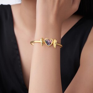 May include: Gold-toned bracelet featuring a heart-shaped locket with a photo, flanked by wing charms. The bracelet has a hinged design and a small gemstone accent. A personalized jewelry item.