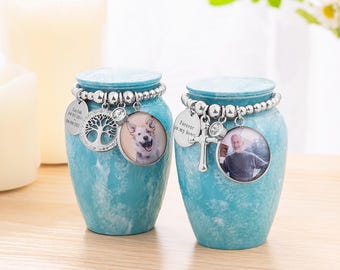 May include: Two turquoise cremation urns with photo pendants. One urn has a photo of a dog, the other a man. Each urn has a beaded bracelet and charms. One charm says "Forever in my heart."