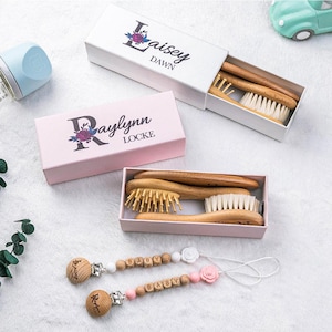 May include: Baby brush and comb sets in white and pink boxes, with the names "Laisey Dawn" and "Raylynn Locke" printed on them. Two pacifier clips with the word "BABY" spelled out in wooden beads are also visible.
