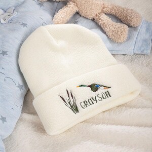 May include: Cream-colored knit beanie with an embroidered duck and cattails design. The name "GRAYSON" is embroidered below the design in green. The beanie is folded at the brim.