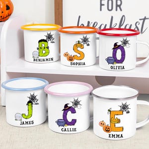 May include: Six white enamel mugs with colored rims, each featuring a large letter decorated with Halloween motifs like pumpkins, spiderwebs, and ghosts. Each mug is personalized with a name printed in black text below the letter.
