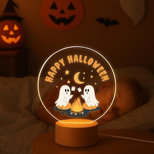 May include: A Halloween night light with a wooden base. The clear, circular light features the words "Happy Halloween" and illustrations of ghosts and a campfire. The light is on, emitting a warm glow.