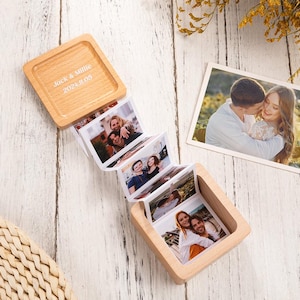 Puede incluir: Una caja de fotos de madera con tapa con bisagras, que muestra una serie de fotografías adjuntas. La tapa está grabada con el texto "Jack & Mille 2024.11.05". La caja está abierta, revelando varias fotos impresas de parejas.