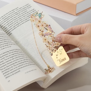 May include: A gold-toned metal bookmark featuring an intricate cutout design of cherry blossoms and butterflies with pastel enamel accents. The bookmark includes a dangling chain with a butterfly charm and is engraved with the name Tina.