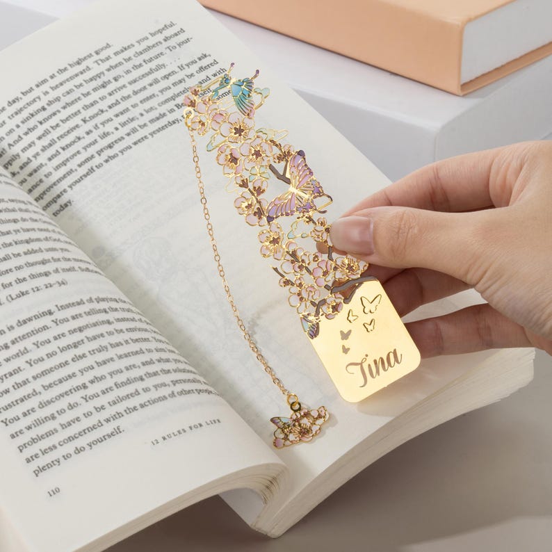 May include: A gold-toned metal bookmark featuring an intricate cutout design of cherry blossoms and butterflies with pastel enamel accents. The bookmark includes a dangling chain with a butterfly charm and is engraved with the name Tina.