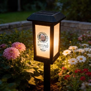 Op de afbeelding: Een zwarte solar tuinlamp op een pin met een rechthoekig paneel van matglas. Het paneel bevat een cirkelvormig portret van een man, de tekst "ALWAYS LOVED NEVER FORGOTTEN", "ALWAYS IN OUR THOUGHTS FOREVER IN OUR HEARTS" en "Thomas 1950-2026".
