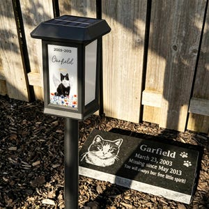 May include: A black solar-powered memorial garden light with a picture of a black and white cat with angel wings, the name "Garfield", and dates 2003-2013. A black granite headstone with a cat portrait and the name "Garfield" is also visible.