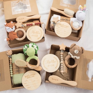 May include: Four baby gift boxes with various items. Each box contains a wooden brush, a crocheted animal, a wooden teether, a pacifier clip, and a wooden disc that says "Hello World". The boxes are brown and filled with baby essentials.