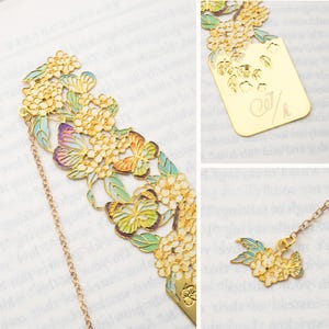 May include: A gold-toned metal bookmark featuring intricate cutouts of flowers and butterflies with enamel accents in shades of blue, purple, and green. A thin gold chain connects to a small floral charm, and the back displays the engraved text CJ/h.