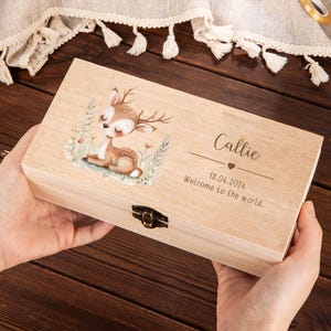 May include: A light wooden box with a deer illustration and the name "Callie" engraved on the lid. The box also includes the date "18.04.2024" and the phrase "Welcome to the world."