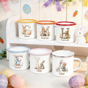 Puede incluir: Tazas de esmalte blanco con bordes coloridos y diseños de conejitos personalizados. Cada taza presenta una letra, un nombre e ilustraciones temáticas de Pascua. Los bordes son naranja, rojo, amarillo, azul, rosa y plateado.