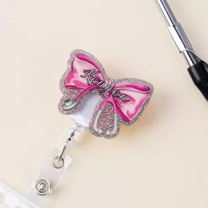 May include: A pink bow-shaped badge reel with the name "Kenyon" in cursive script. The bow and stethoscope design are outlined with glitter. The reel has a clear plastic strap and a silver metal clip.