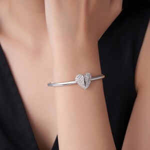 May include: A silver-toned bracelet featuring a heart-shaped design. The heart is split into two halves and encrusted with small, sparkling stones. The bracelet is worn on a wrist, showcasing its elegant and delicate appearance.