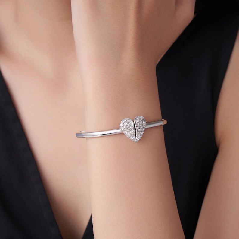 May include: A silver-toned bracelet featuring a heart-shaped design. The heart is split into two halves and encrusted with small, sparkling stones. The bracelet is worn on a wrist, showcasing its elegant and delicate appearance.