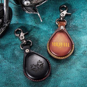 Puede incluir: Dos llaveros de cuero con cierres metálicos. Uno es negro con un gráfico de motocicleta y las palabras "MOTO CLUB". El otro es marrón con el nombre "MICHAEL" en letras doradas. Ambos tienen forma de lágrima.