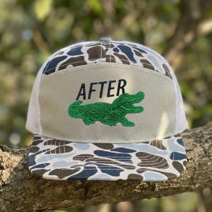 May include: A camouflage-patterned trucker hat with a white mesh back and a khaki front panel. The front panel features the word "AFTER" above a green alligator design.