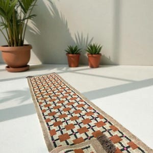 May include: A rectangular runner rug with a geometric pattern of orange, black, and pink squares on a cream background. The rug has a tan border and fringe on the end. The rug is displayed on a white surface.