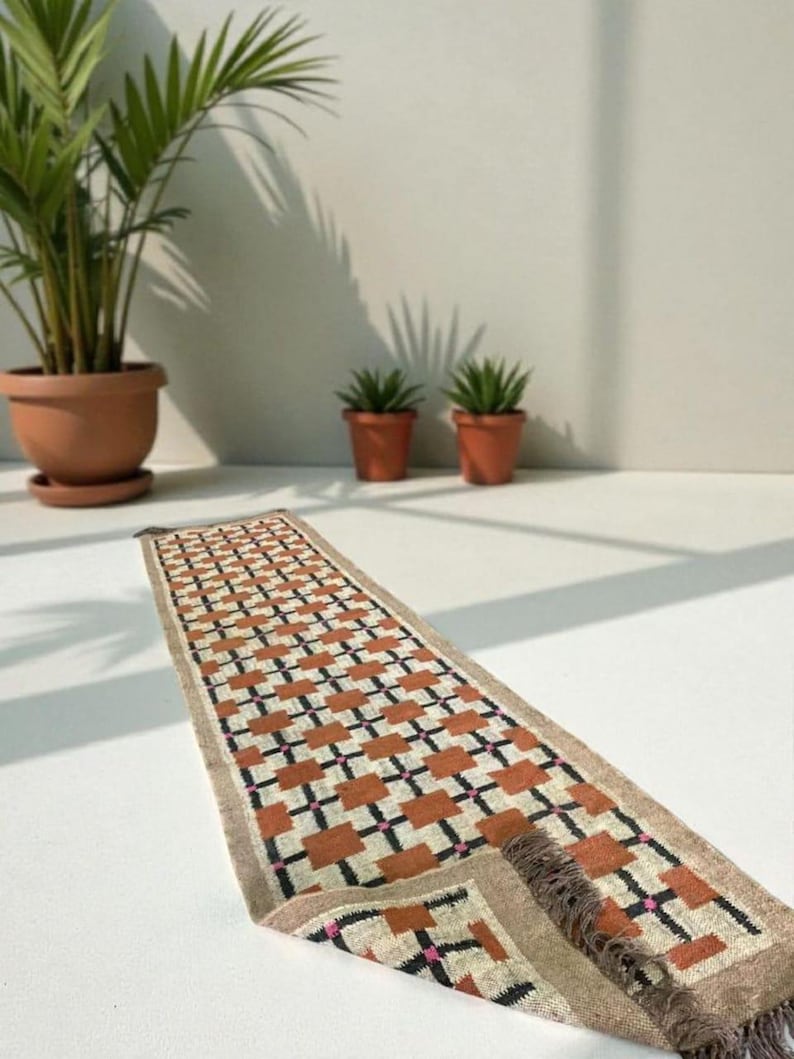 May include: A rectangular runner rug with a geometric pattern of orange, black, and pink squares on a cream background. The rug has a tan border and fringe on the end. The rug is displayed on a white surface.