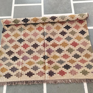 May include: A rolled-up, rectangular rug with a repeating geometric pattern. The pattern features multi-colored cross-like shapes in shades of brown, red, green, and purple on a beige background. The rug has a fringed edge.