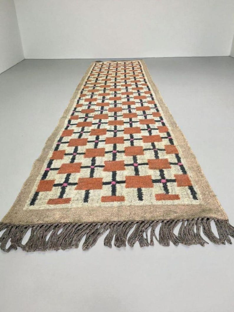 May include: A long, rectangular rug with a geometric pattern. The rug features a repeating design of orange rectangles, black crosses, and pink dots on a cream background. The rug has a tan border and fringe.
