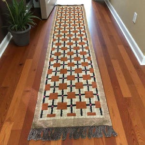 May include: A rectangular runner rug with a geometric pattern of brown squares and black cross shapes on a beige background. The rug has a brown fringe on both ends. The rug is placed on a hardwood floor.