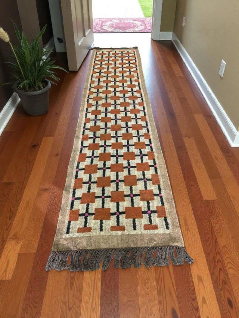 May include: A rectangular runner rug with a geometric pattern of brown squares and black cross shapes on a beige background. The rug has a brown fringe on both ends. The rug is placed on a hardwood floor.