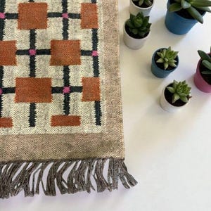 May include: A rectangular rug with a geometric pattern of brown squares, black lines, and pink accents. The rug has a beige border and fringe. Small potted succulents in white, blue, and pink pots are arranged on a white surface.