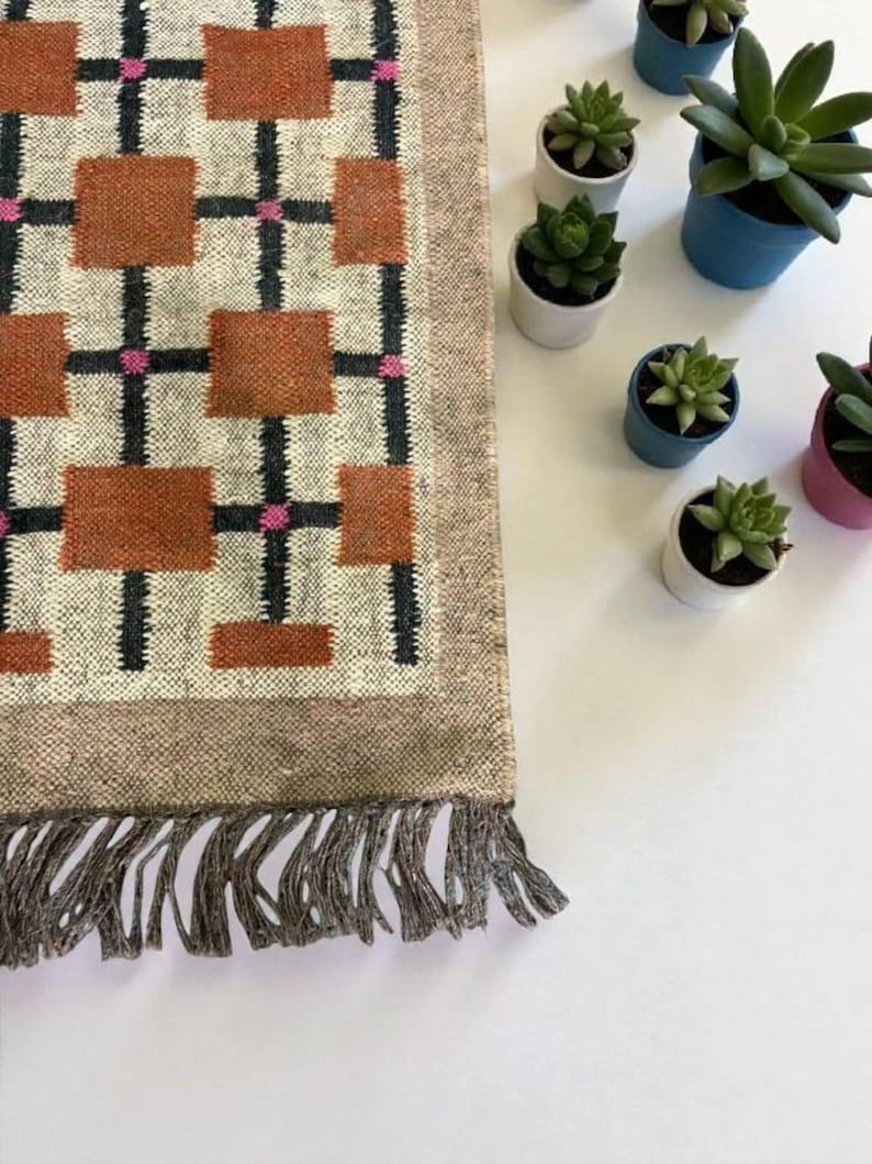 May include: A rectangular rug with a geometric pattern of brown squares, black lines, and pink accents. The rug has a beige border and fringe. Small potted succulents in white, blue, and pink pots are arranged on a white surface.