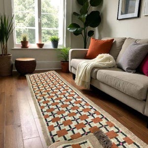 May include: A long, rectangular rug with a geometric pattern of orange, black, and pink squares on a cream background. The rug is placed on a hardwood floor in a living room setting, with a sofa and plants visible.