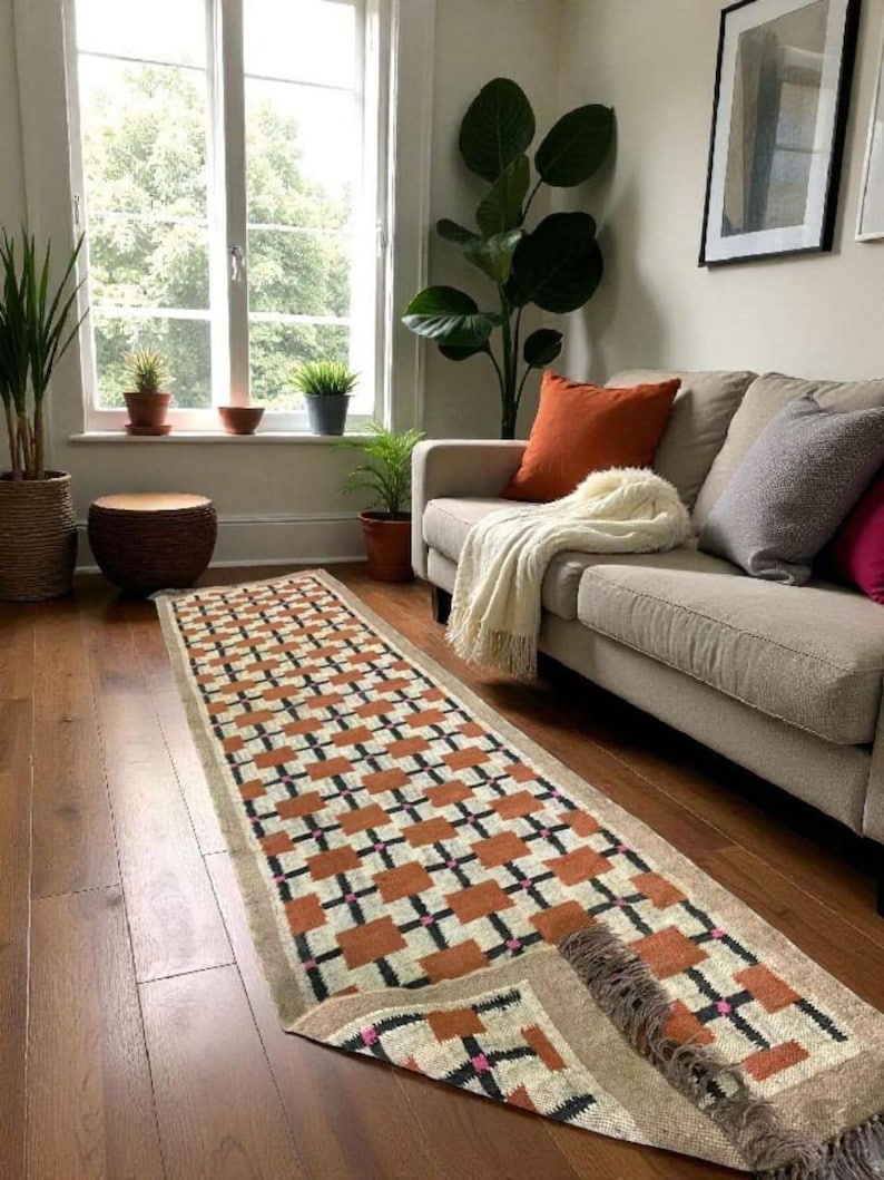 May include: A long, rectangular rug with a geometric pattern of orange, black, and pink squares on a cream background. The rug is placed on a hardwood floor in a living room setting, with a sofa and plants visible.