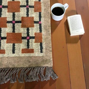 May include: A rectangular rug with a geometric pattern of brown squares, black lines, and pink accents. The rug has a fringed edge. A white mug filled with black coffee and an open book are next to the rug on a wooden surface.