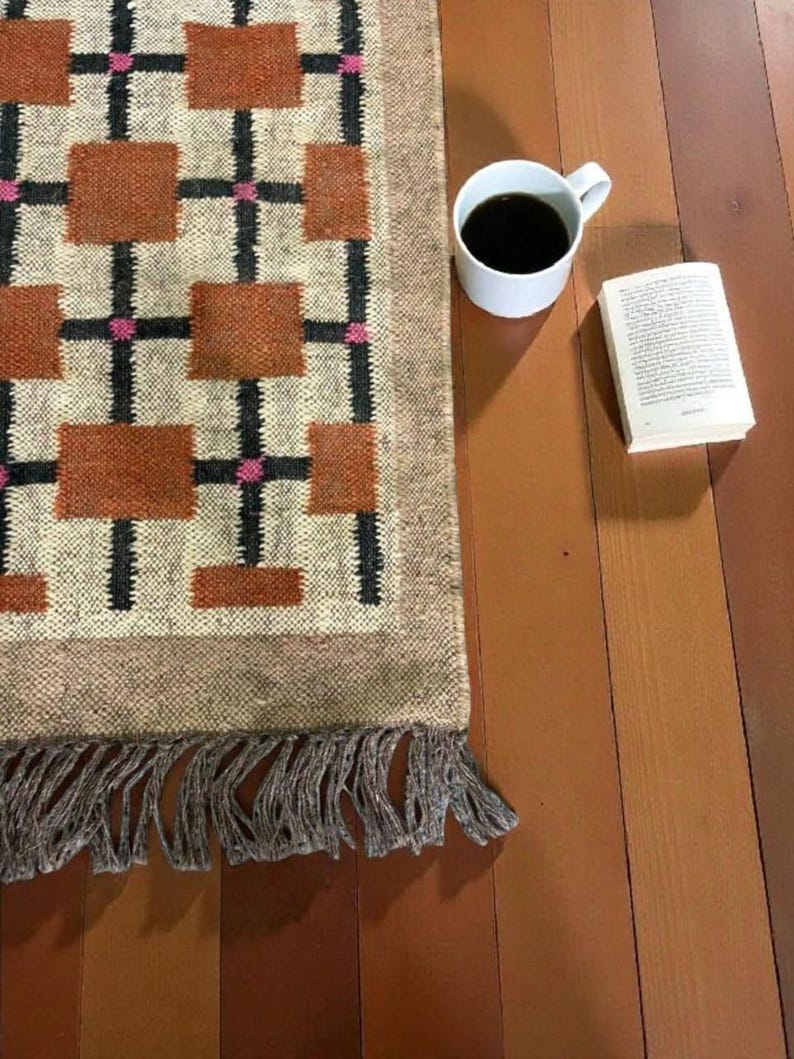 May include: A rectangular rug with a geometric pattern of brown squares, black lines, and pink accents. The rug has a fringed edge. A white mug filled with black coffee and an open book are next to the rug on a wooden surface.